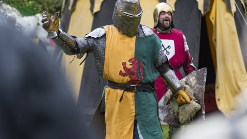A medieval knight with a helmet on holding his arm in a victory pose while another knight laughs behind him. There are tents in the background.